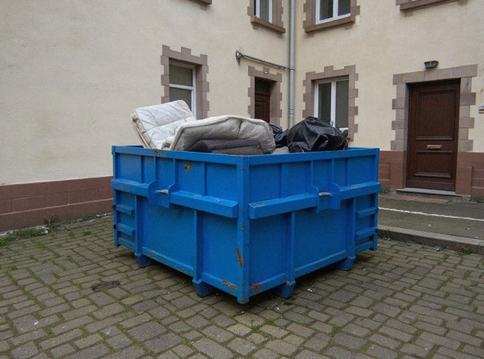 Benne bleue chargée de matelas, d'un canapé et de sacs d'encombrants dans une cour de Haguenau, location de benne pour un débarras